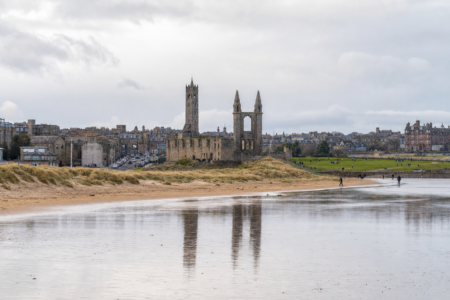 Ruins of a historic cathedral with reflections on a wet sandy beach, surrounded by a coastal townscape.