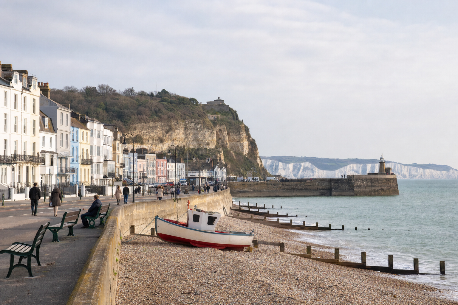 A pebbled beach with a small boat, a promenade with benches, cliffside houses, and a distant lighthouse under a cloudy sky.