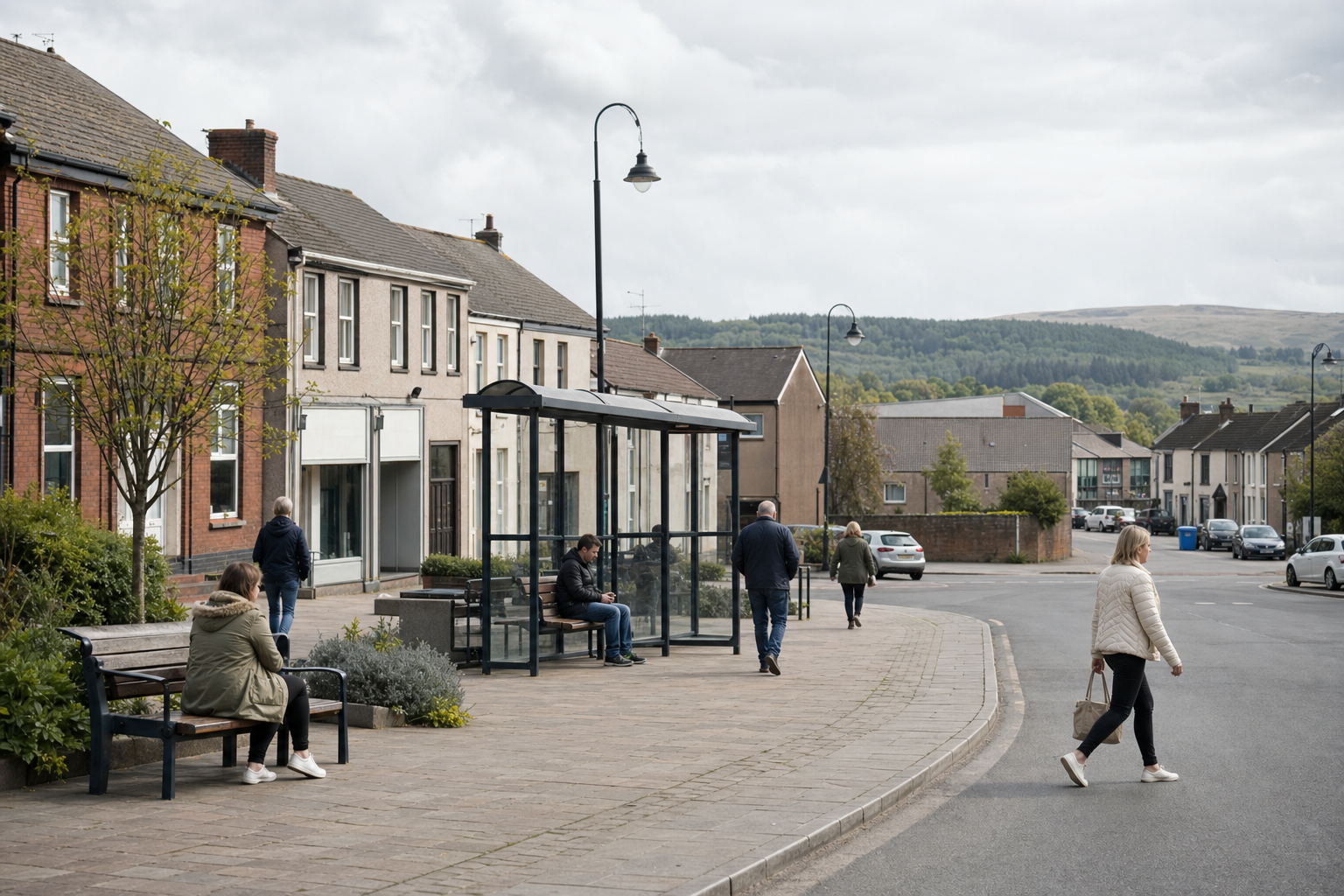 People wait at a bus shelter on a quiet street in a small town, with hills in the background.