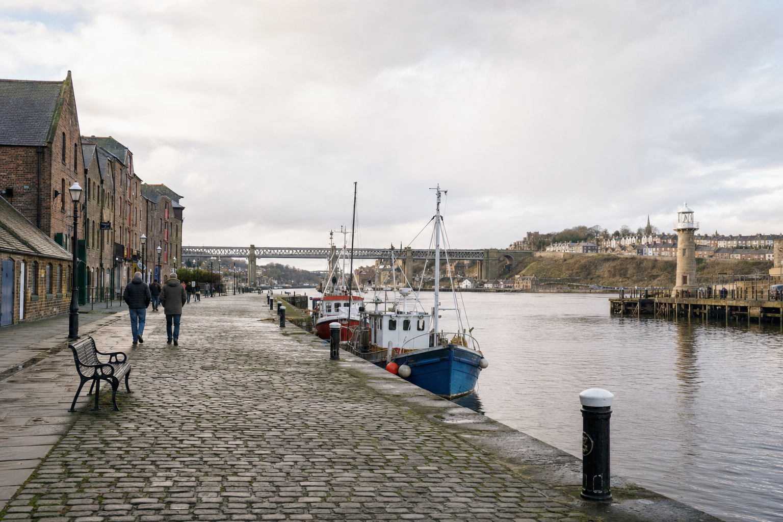 A riverside walkway with docked boats, people walking, and a bridge in the background.