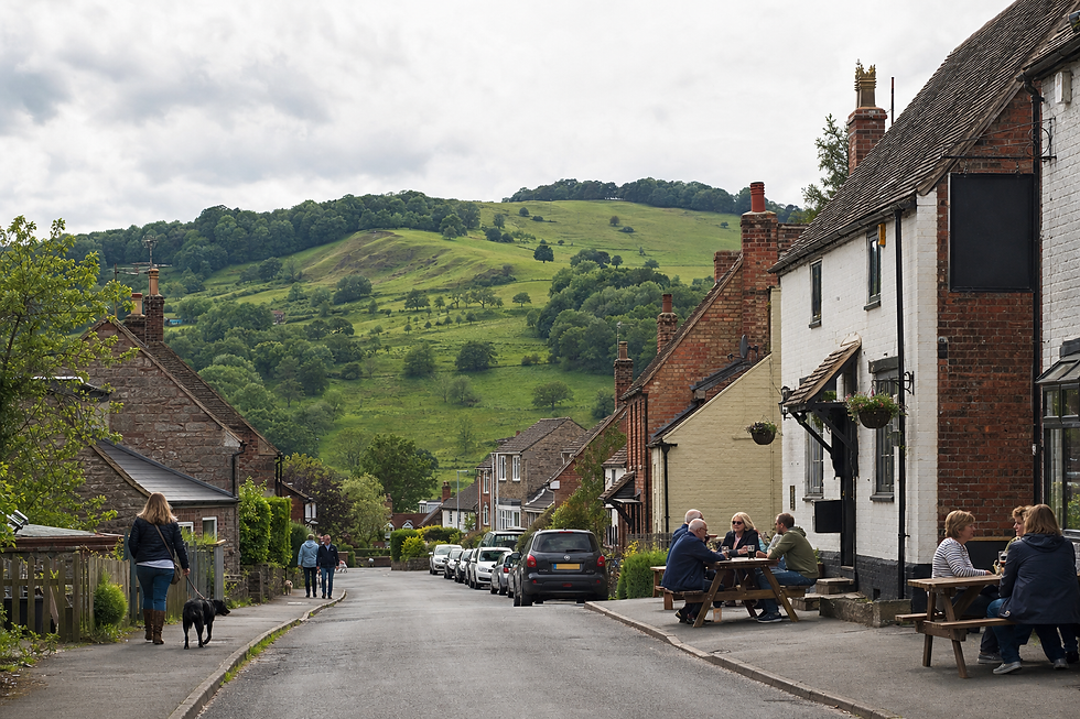 People sitting at outdoor tables of a pub in a quaint village with lush green hills in the background.