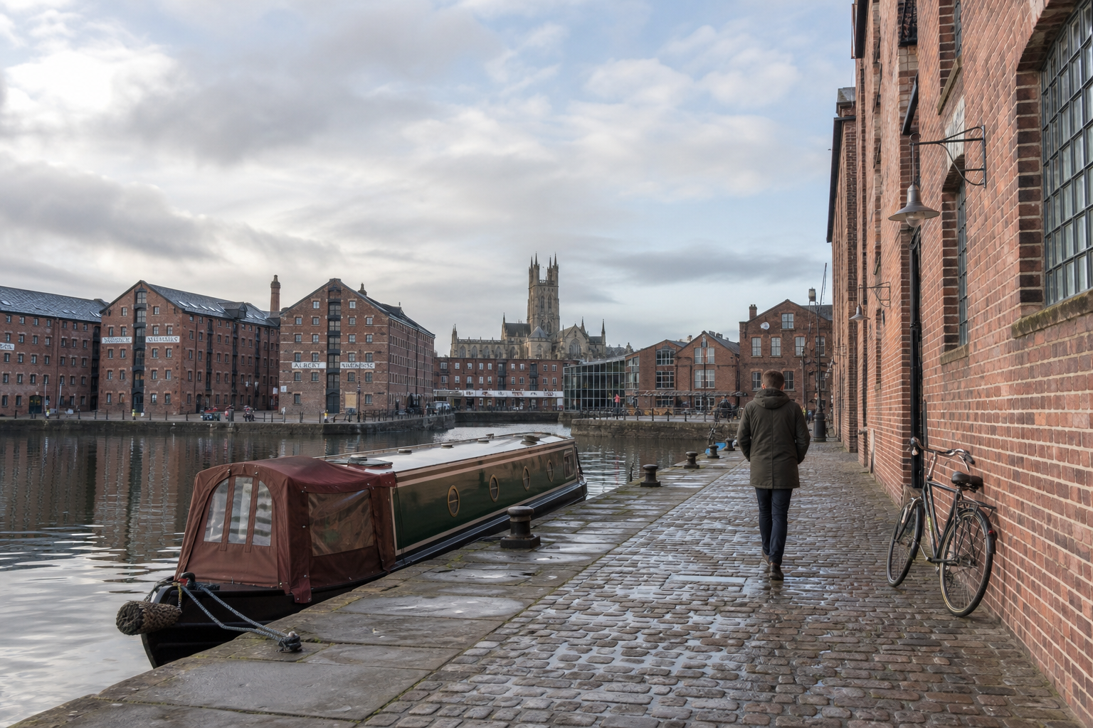 A person walks along a cobbled towpath beside a canal boat and red brick warehouses, with a cathedral in the background.
