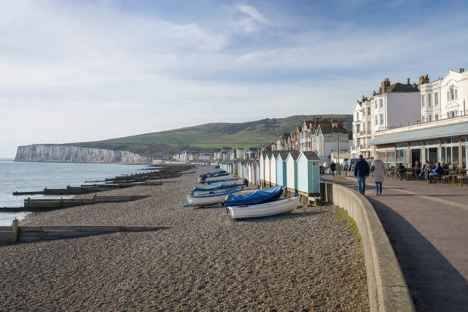 Coastal promenade with colourful beach huts, boats on a pebble beach, and cliffs in the background under a blue sky.