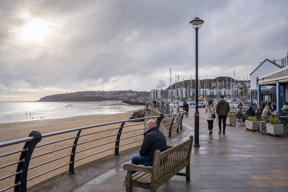 People strolling along a seaside promenade near a marina on a cloudy day, with a man seated on a bench.