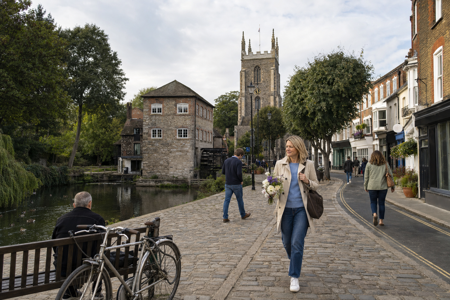 A woman carrying flowers strolls along a cobbled street beside a historic stone building and a church tower.