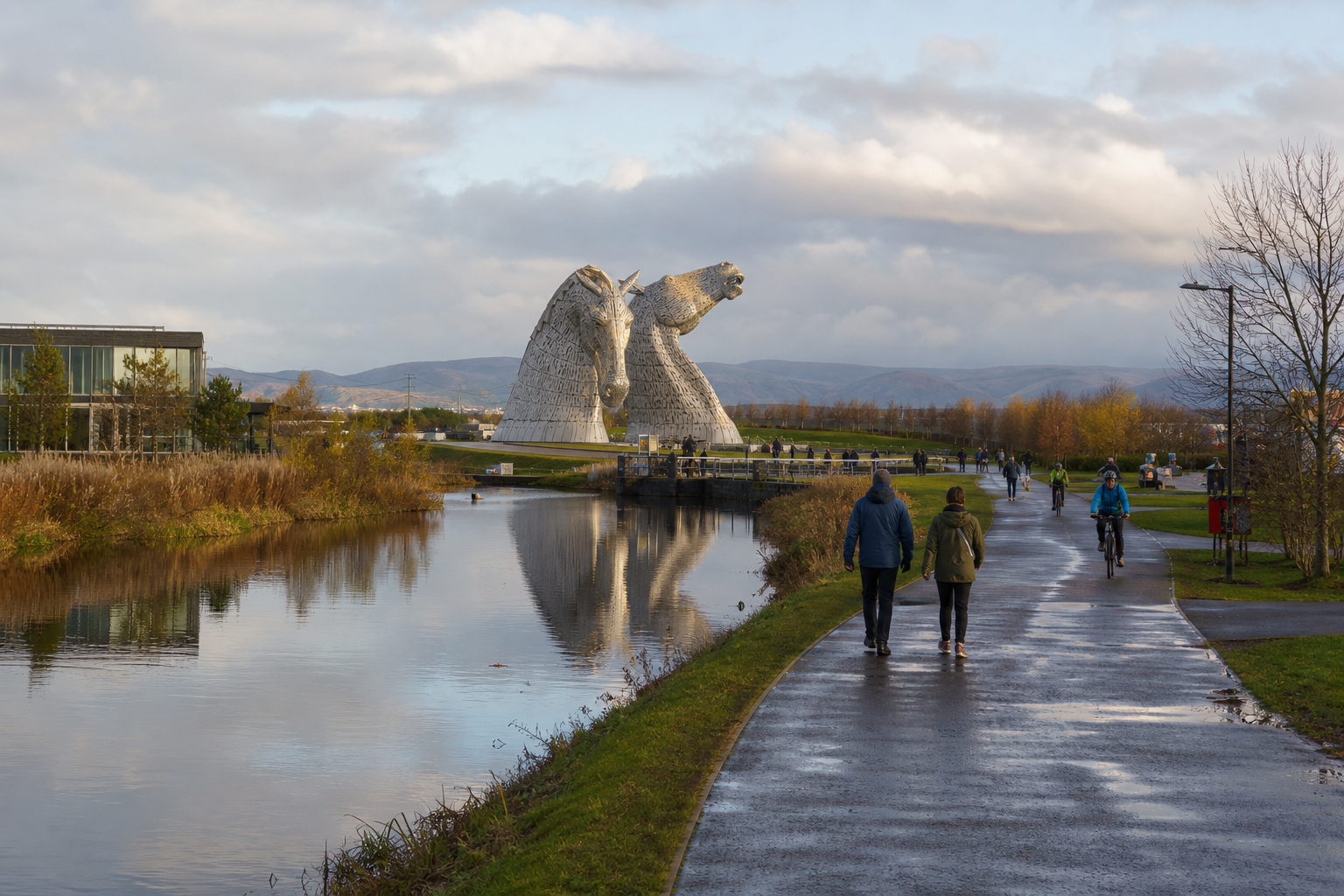 People walking and cycling near large horsehead sculptures by a canal under a cloudy sky.