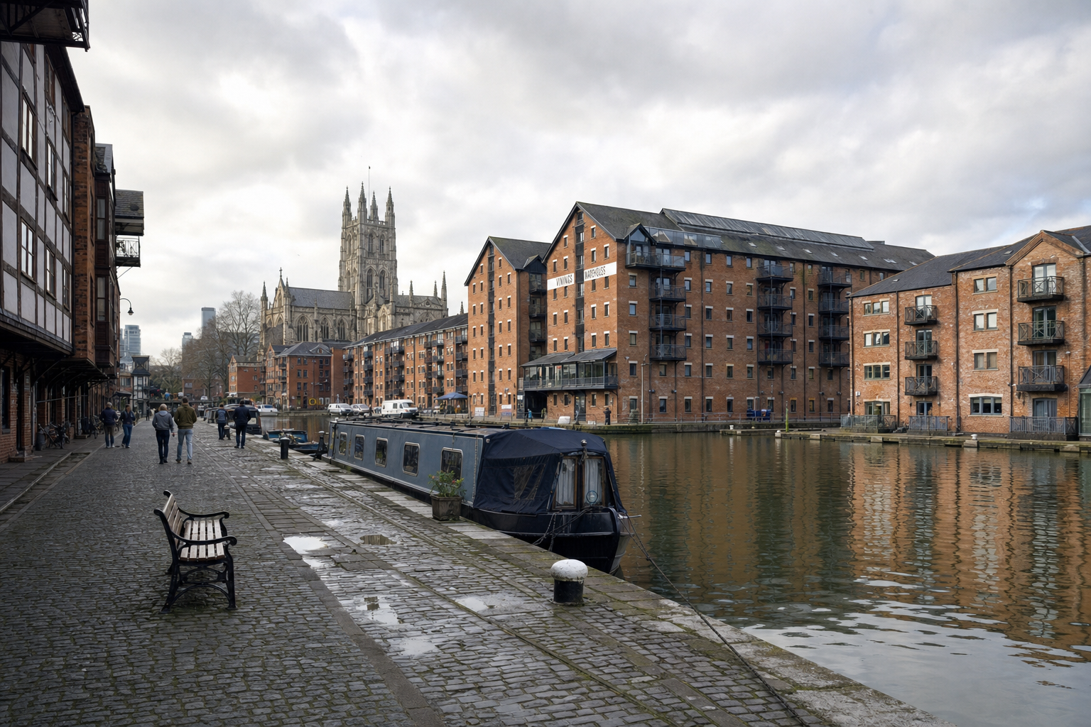 Canal scene with a narrowboat moored alongside red brick buildings and a distant cathedral under a cloudy sky.
