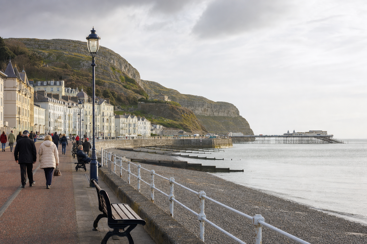 People walking along a seaside promenade with a view of a pier and coastal cliffs in the background.