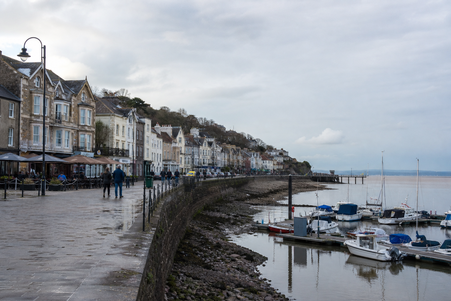 Coastal promenade with Victorian houses and moored boats under a cloudy sky.
