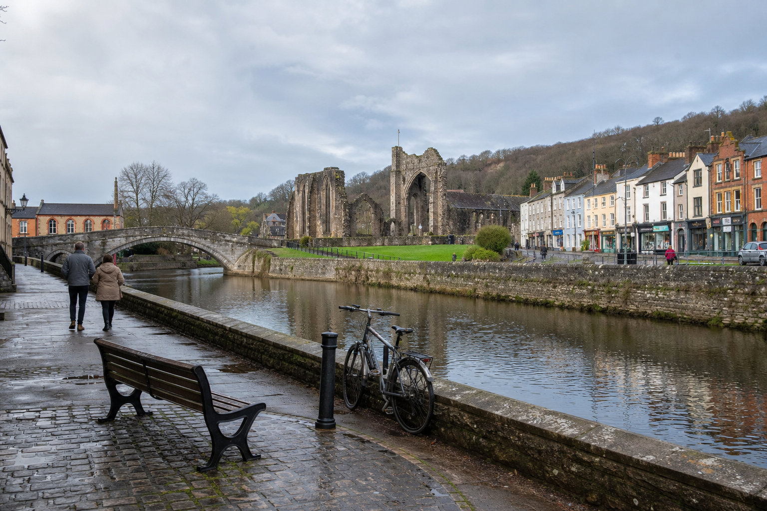 A couple walks along a riverside path near a stone bridge and historic ruins, with colourful buildings lining the opposite bank.