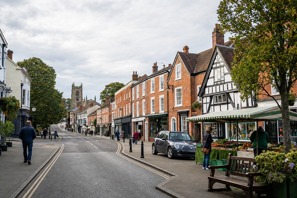 Quaint British high street with red-brick buildings, a market stall, and a church tower in the distance.