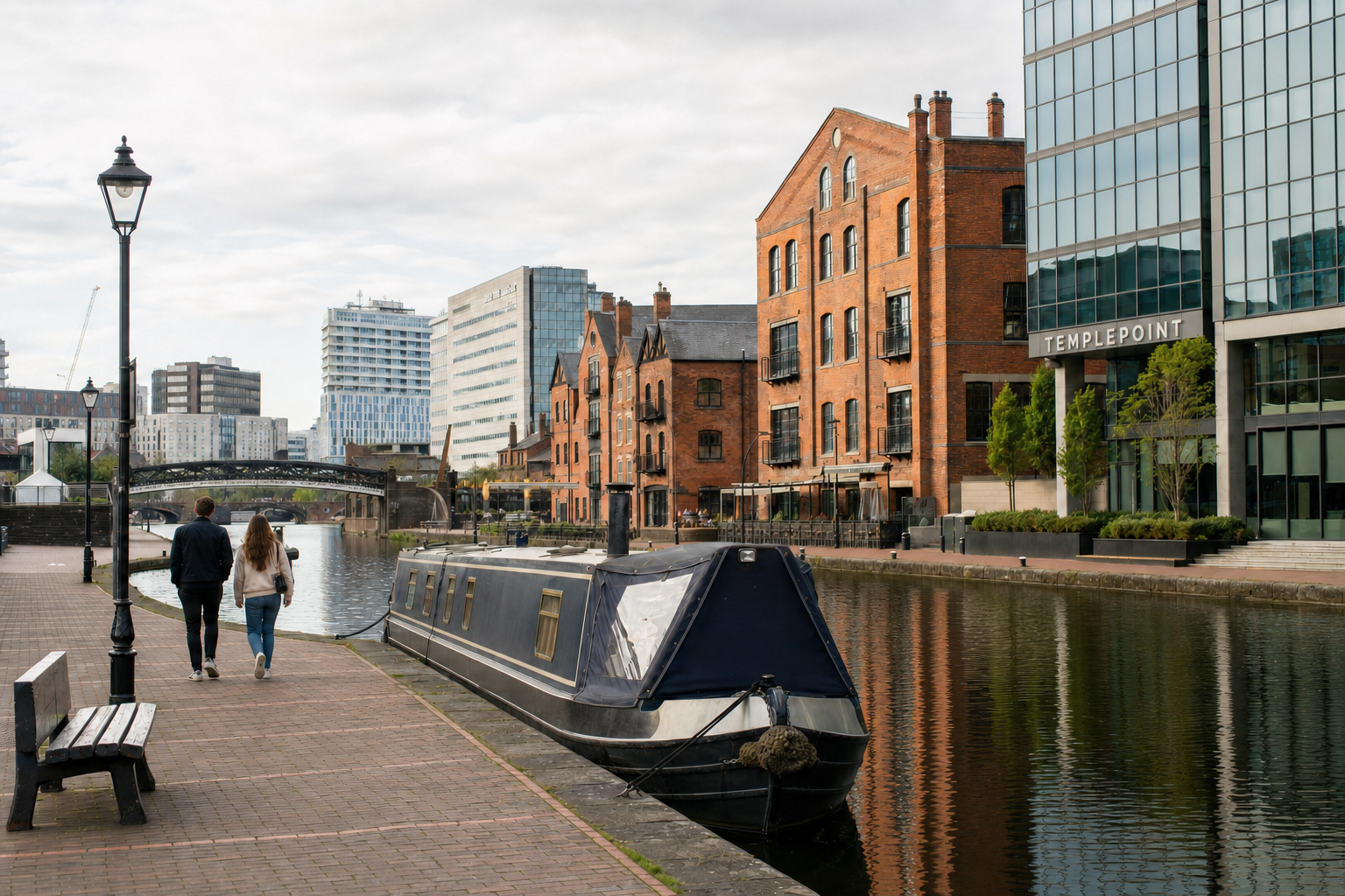A narrowboat moored on a canal with people walking along a towpath lined by red brick buildings and modern structures.