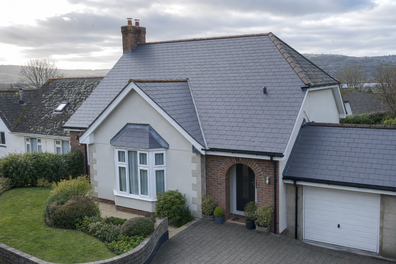 A detached house with a grey slate roof, brick chimney, and a neatly landscaped front garden under a cloudy sky.