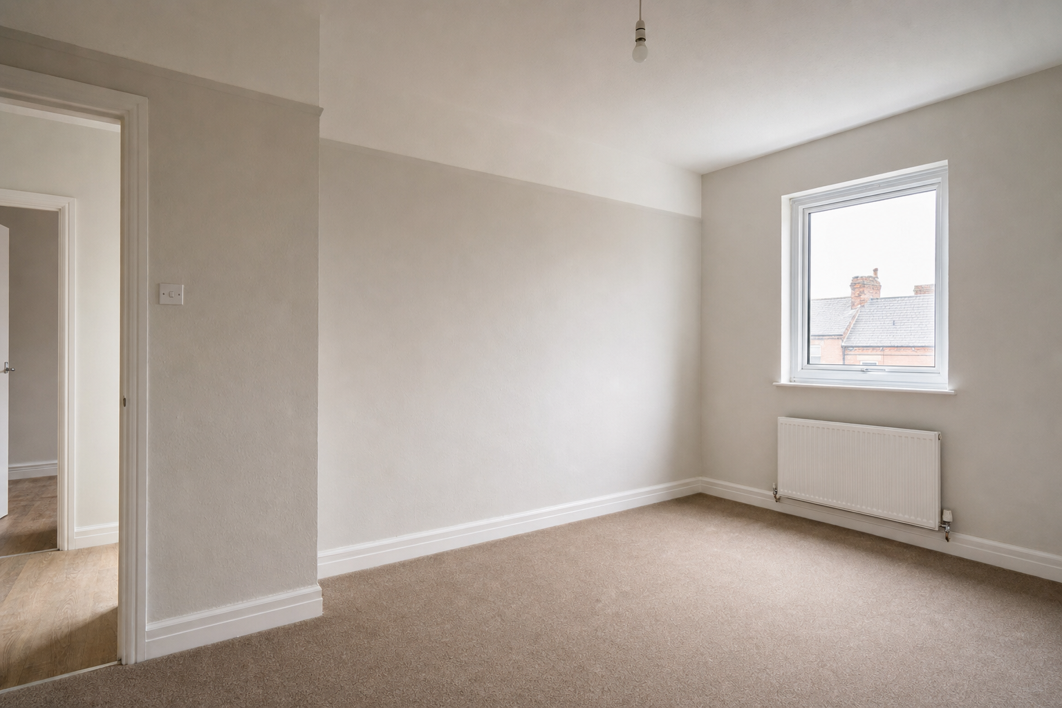 Empty room with beige carpet, white walls, a window showing rooftops, and a radiator beneath the window.