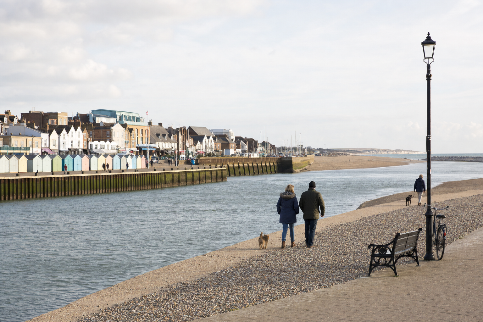 People walking dogs along a riverbank with colourful beach huts and coastal houses in the background.