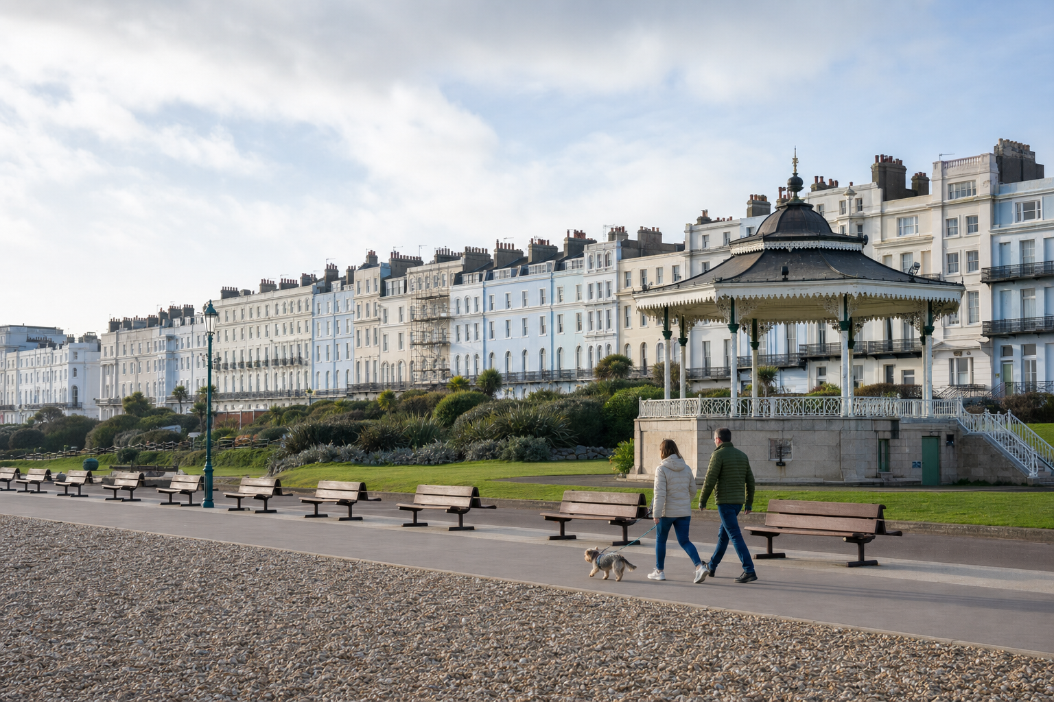 A couple walks a dog along a seaside promenade near a bandstand and Victorian terraced houses.