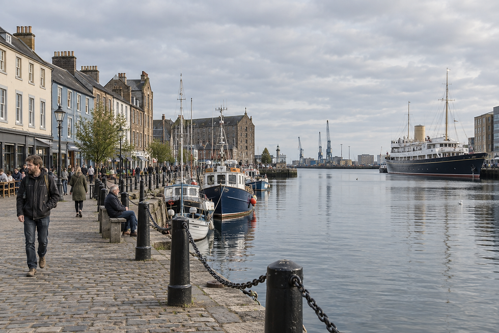 Waterfront promenade with people walking, moored boats, and historic buildings along a harbour under a cloudy sky.