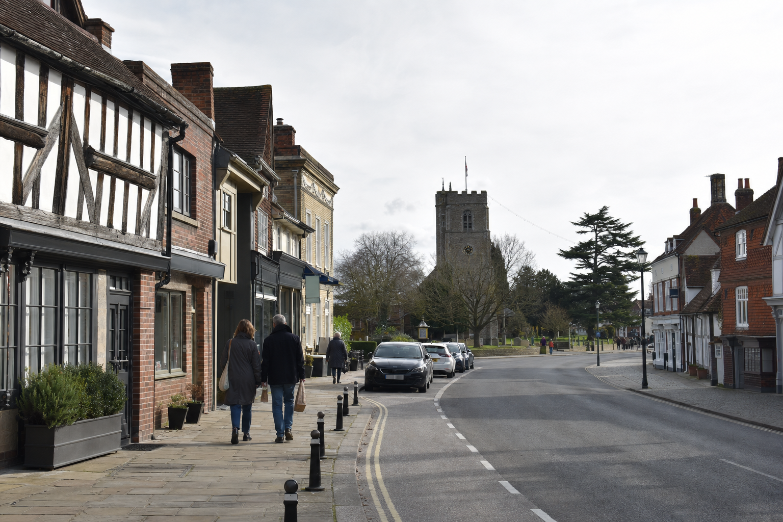 Historic street scene with people walking past timber-framed buildings and a church tower in the background.