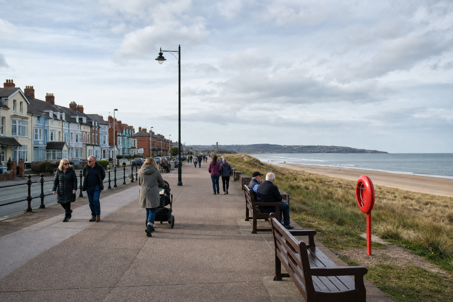 People walking along a coastal promenade with colourful houses and benches overlooking the beach.
