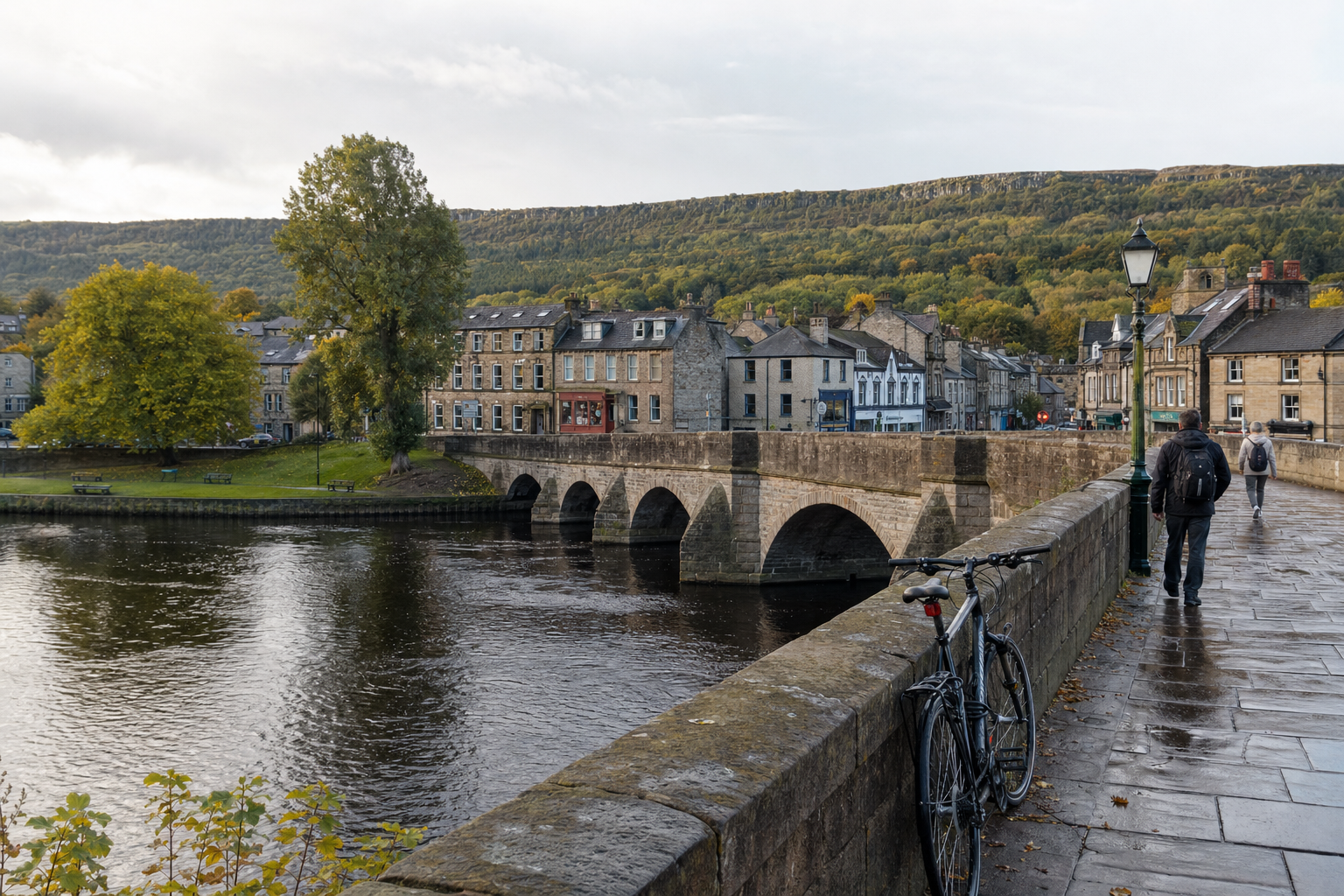 A stone bridge spans a river with nearby buildings and two people walking on its path, set against a backdrop of green hills.