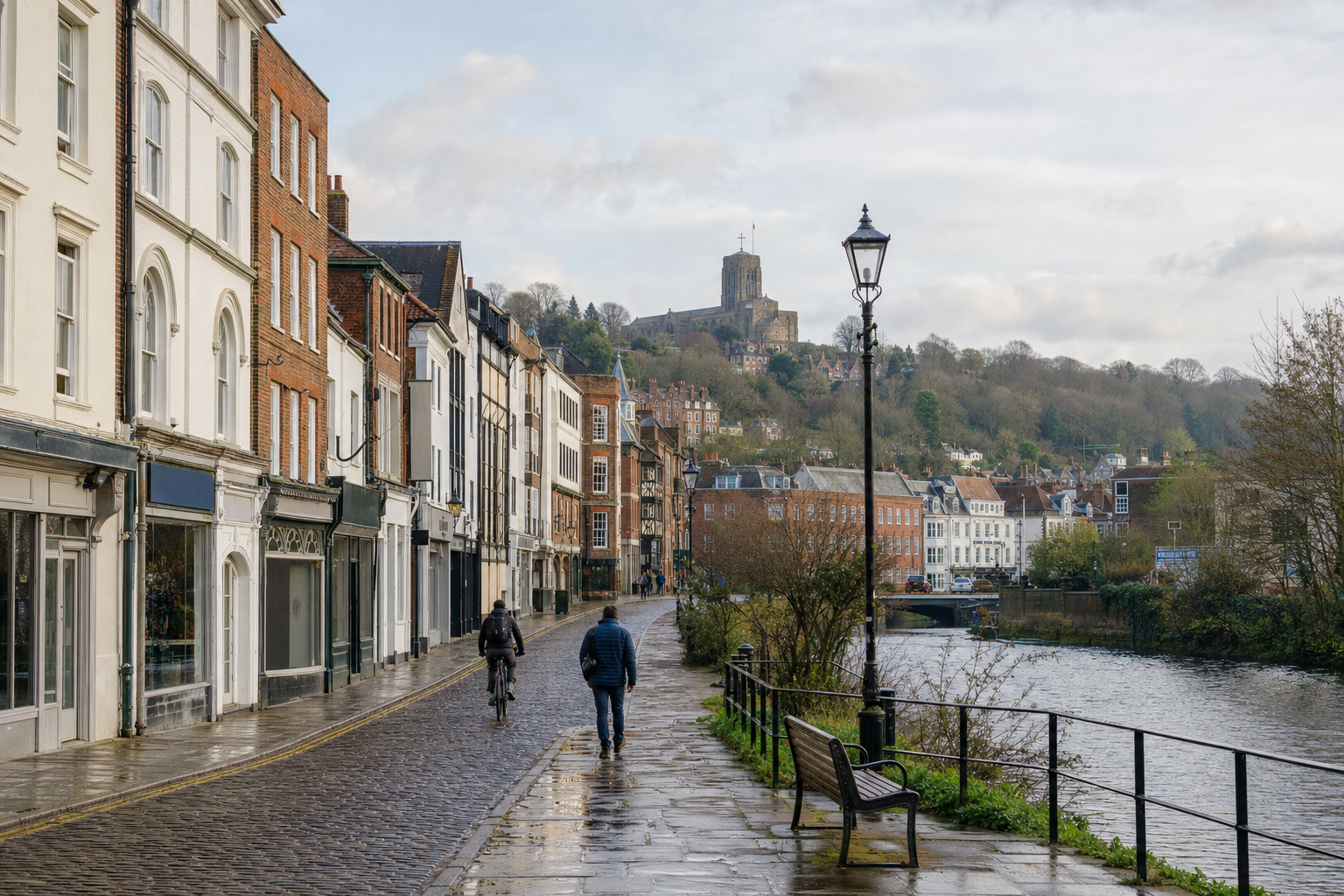 A cobblestone street lined with historic buildings beside a river, with a castle on a hill in the background.