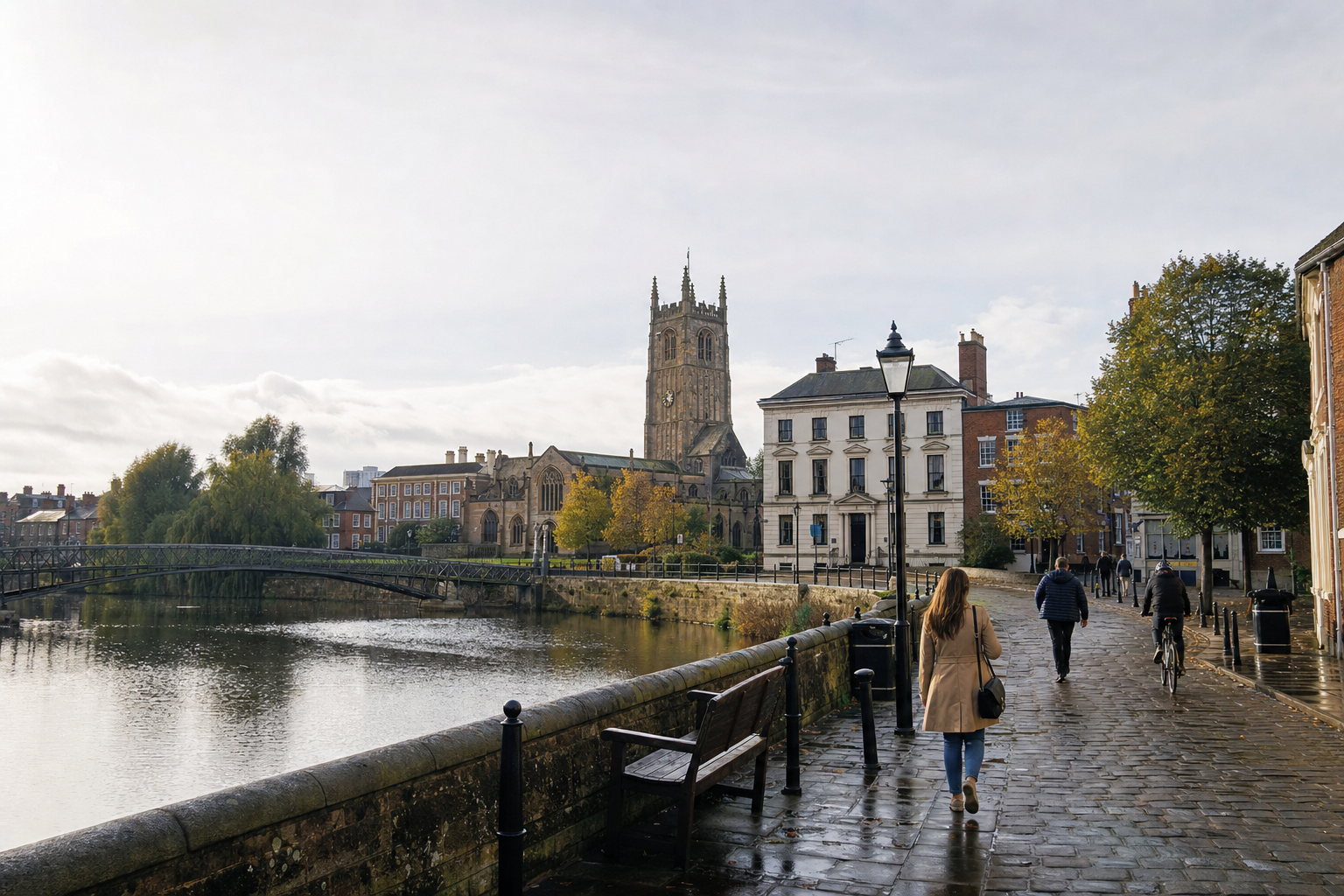 People walking along a riverside path with historic buildings and a church in the background.