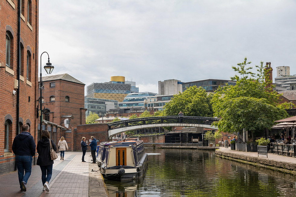 People stroll along a canal in an urban setting with modern and historic buildings in the background.