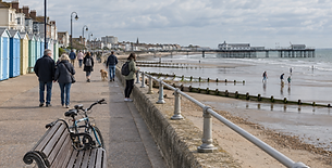 People walk along a seaside promenade with colourful beach huts and a pier in the background.