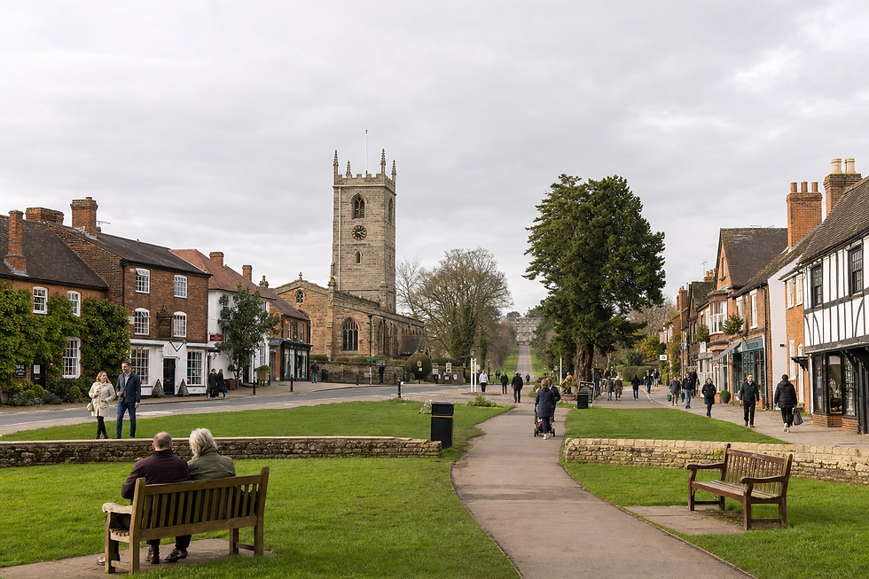 A bustling village scene with people walking near a historic church and red-brick buildings under a cloudy sky.