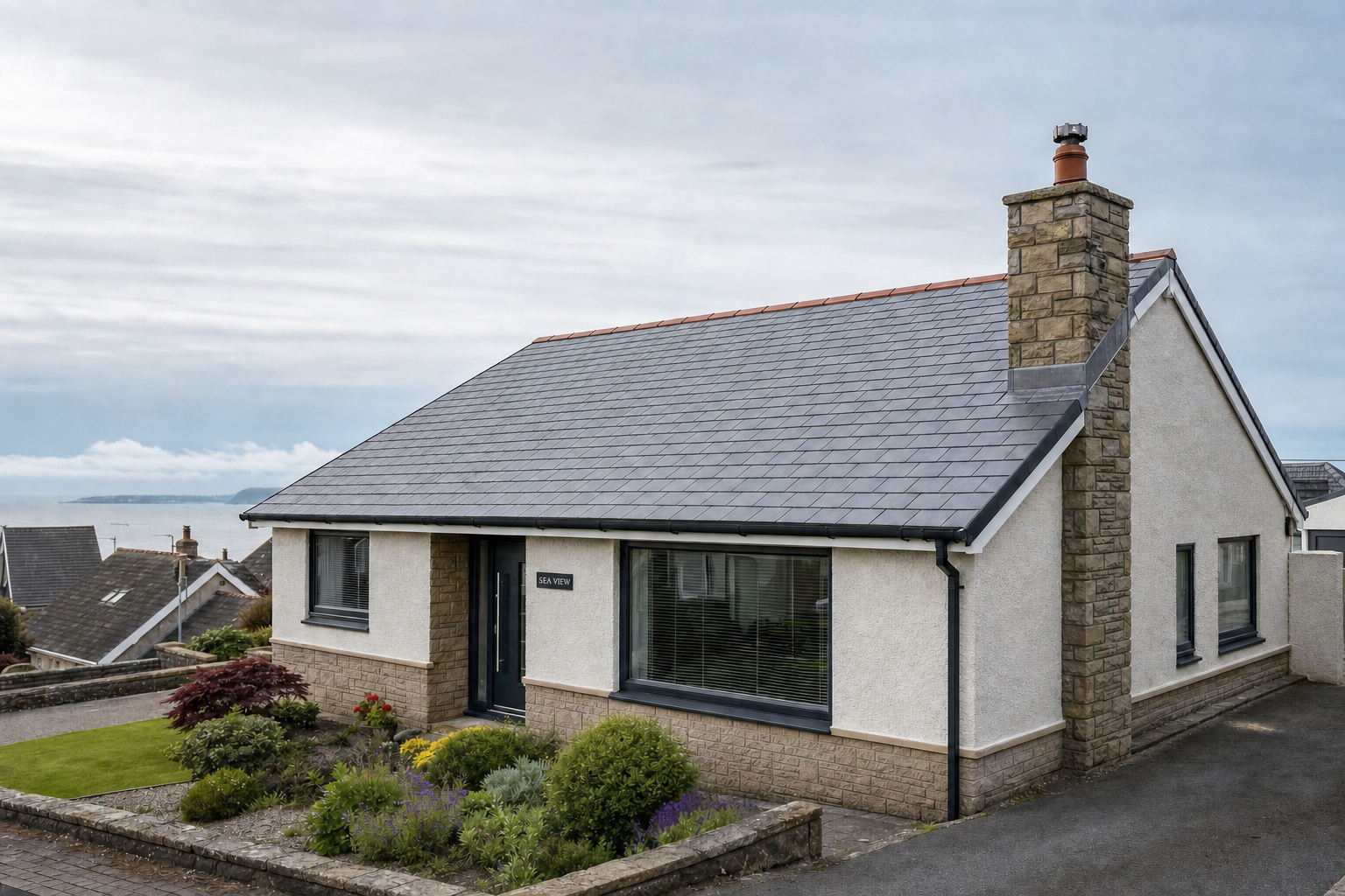 Bungalow with stone chimney and manicured garden, overlooking a seaside view under a cloudy sky.