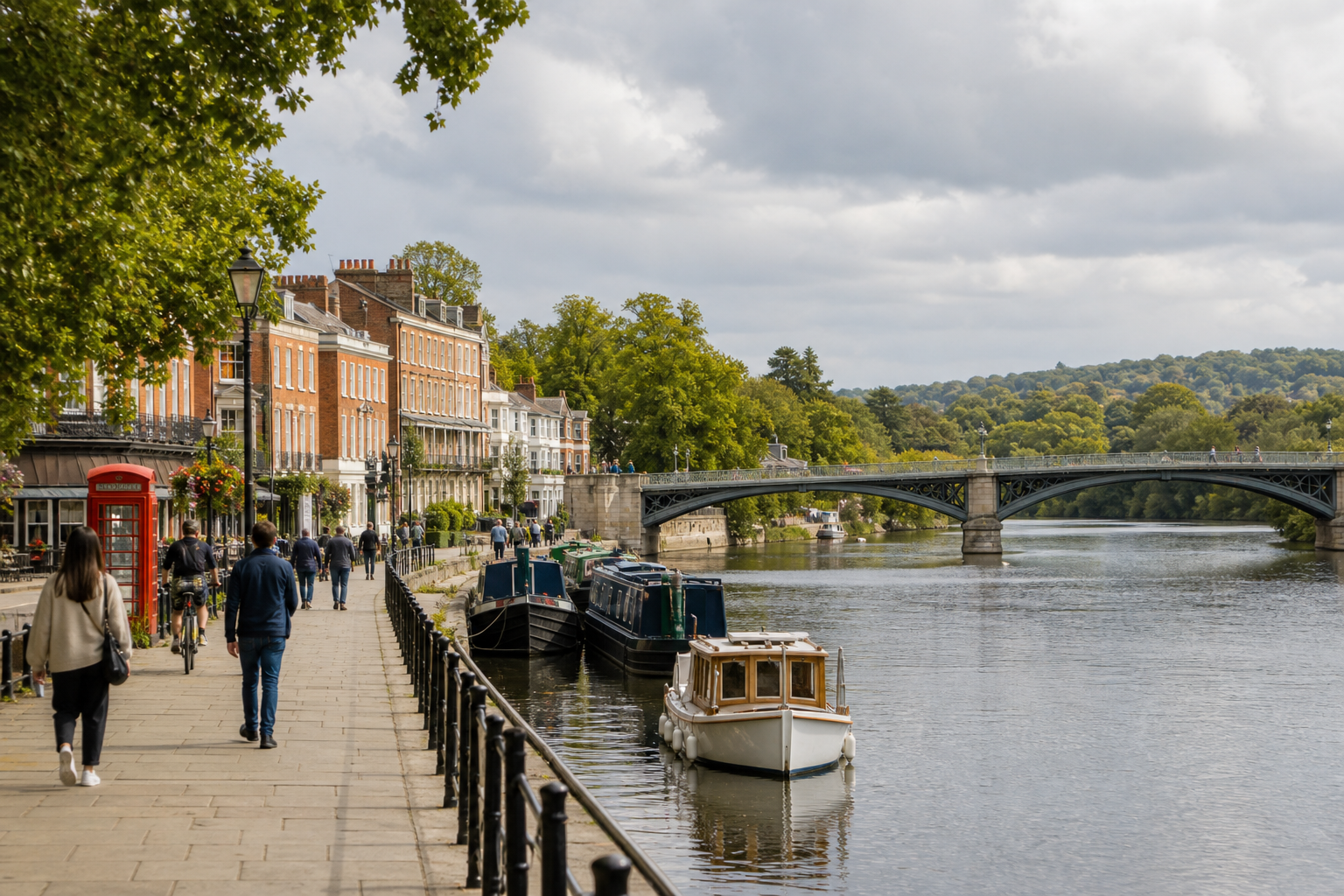 Riverside path with people walking, next to boats and historic buildings under a cloudy sky.