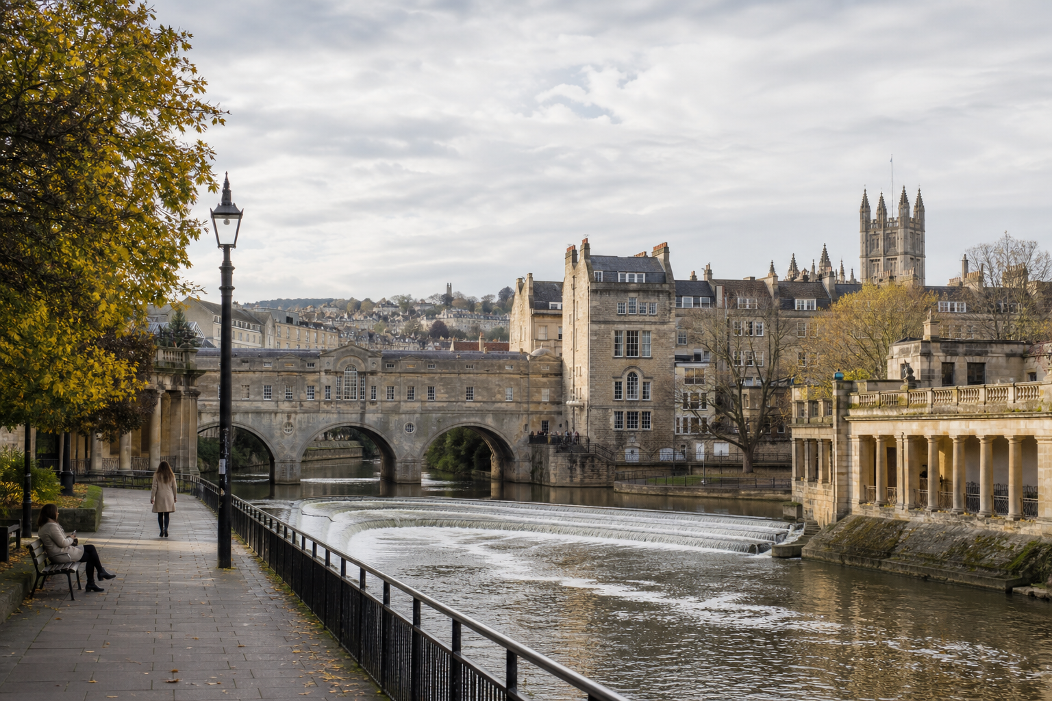 The historic Pulteney Bridge in Bath spans the River Avon under a cloudy sky, with autumn trees lining the riverside path.