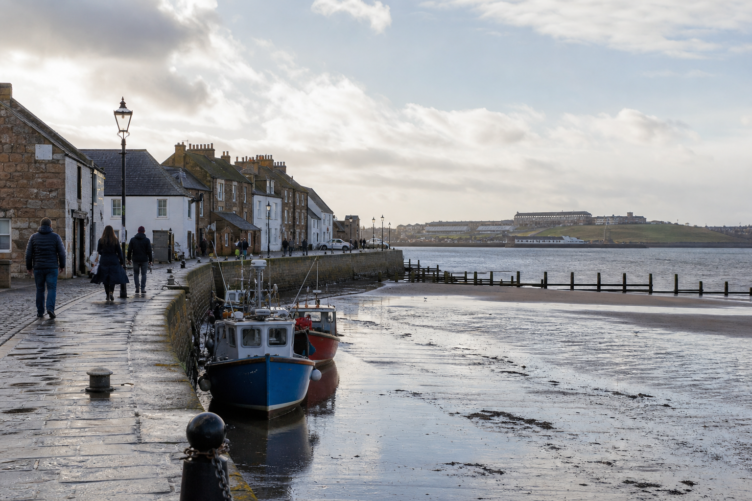 A coastal walkway with people strolling alongside boats docked by stone houses, under a cloudy sky near a waterfront.