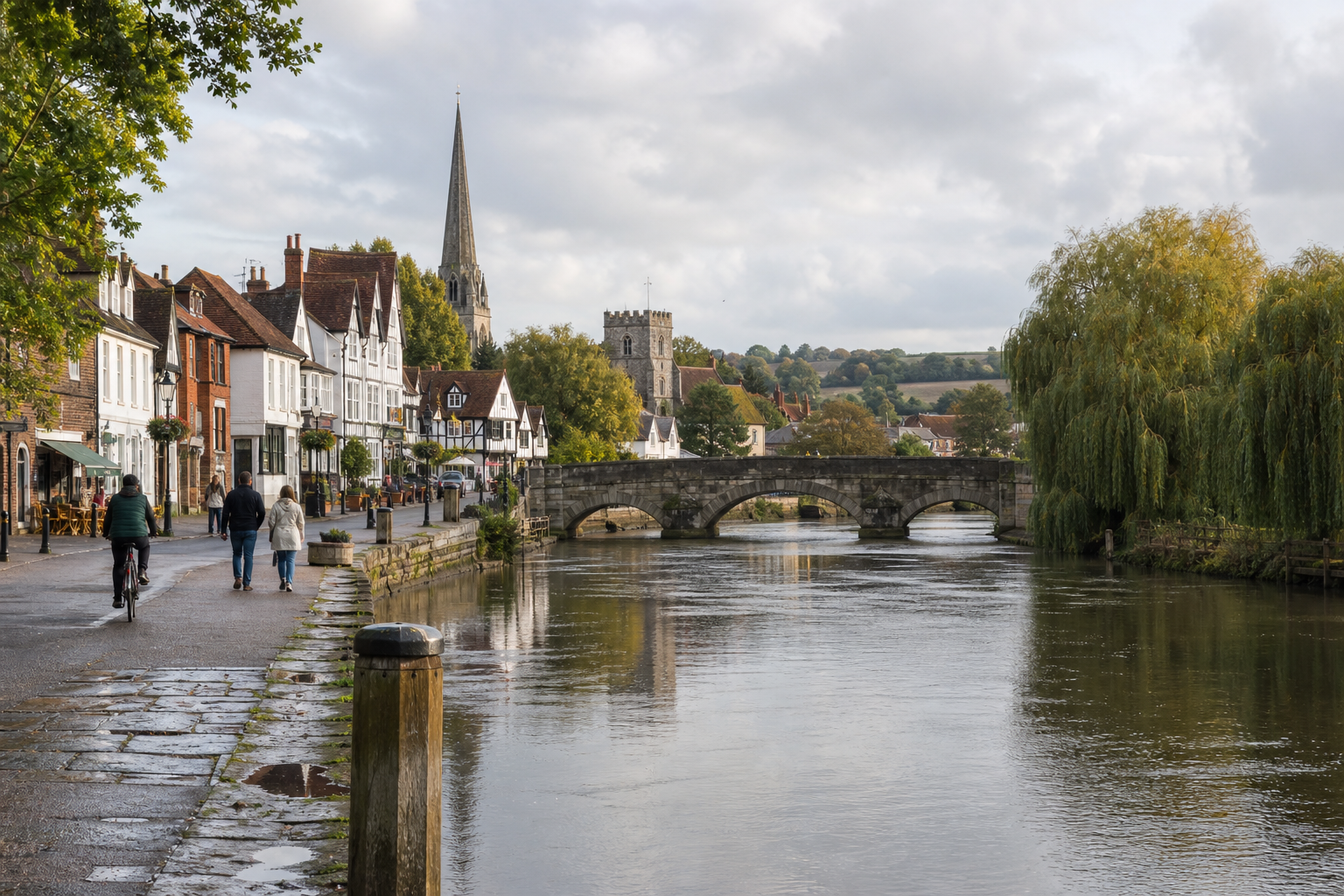 Quaint riverside town with people walking along the path, a cyclist, and a stone bridge across the river.