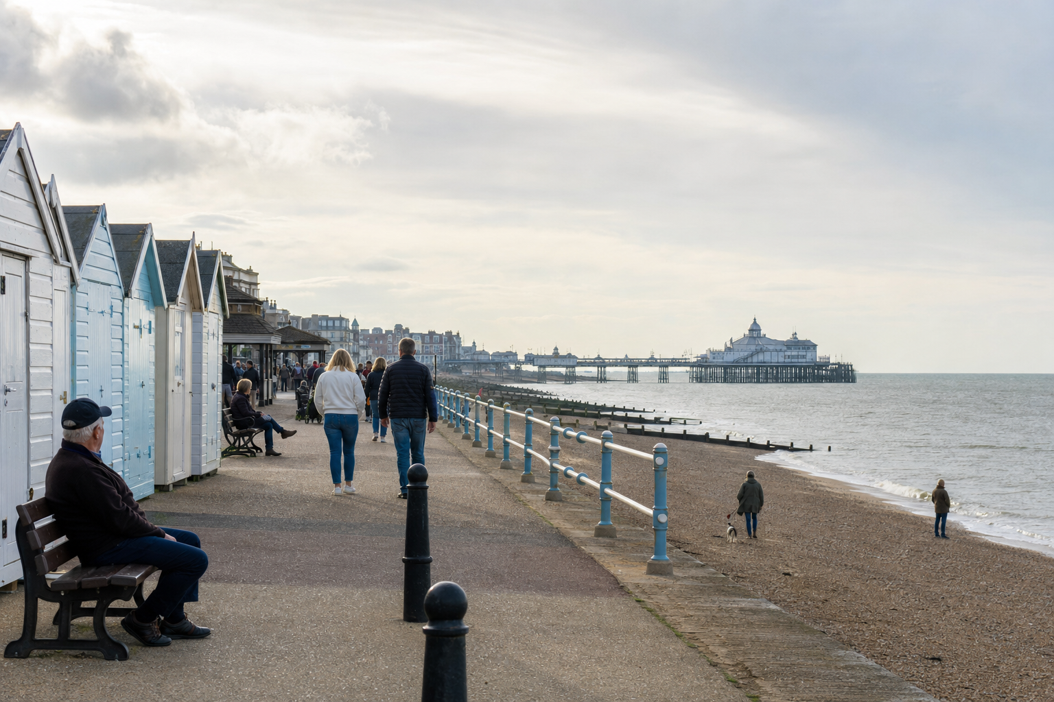 People strolling along a seaside promenade with colourful beach huts and a distant pier.