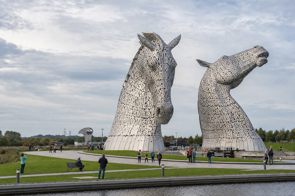 Large horse head sculptures, known as the Kelpies, with visitors walking nearby on a sunny day.