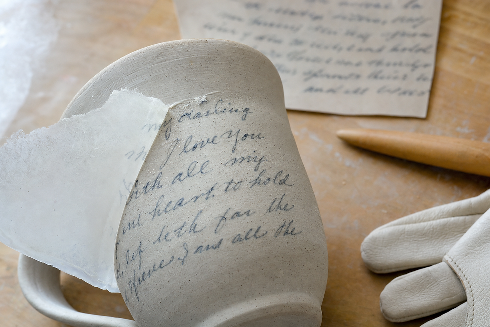 Clay mug with handwritten text being transferred, next to gloves and a wooden tool on a workbench.