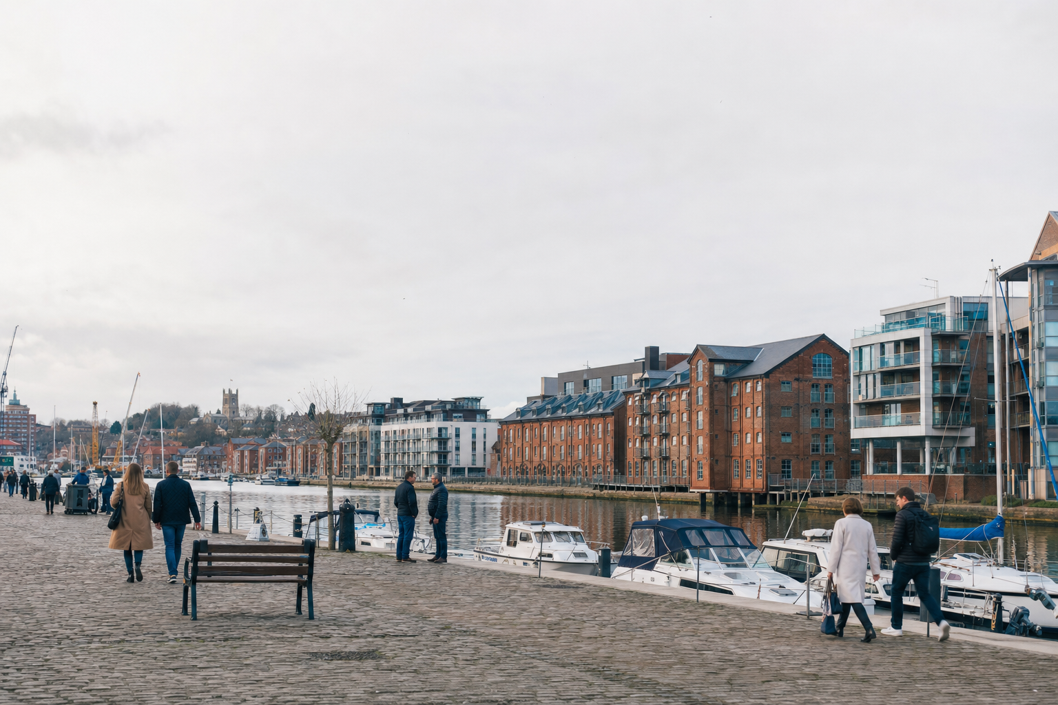 People walking along a cobblestone quay with moored boats and historic buildings in the background.