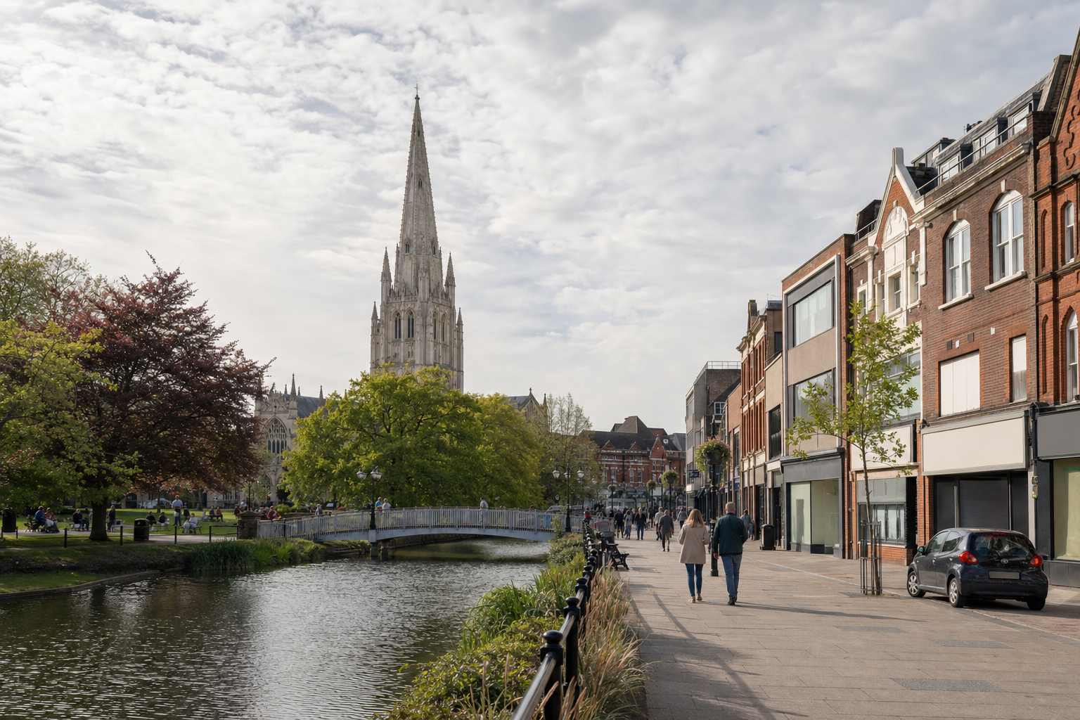 Pedestrian walkway along a canal with people, trees, and a prominent church spire in the background.