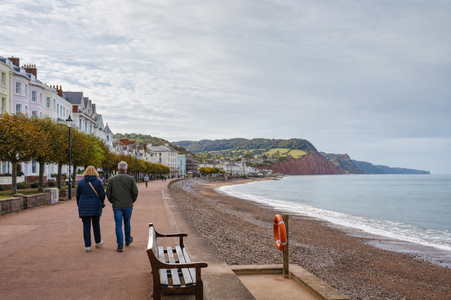 A couple strolls along a seaside promenade with colourful houses and cliffs in the background.