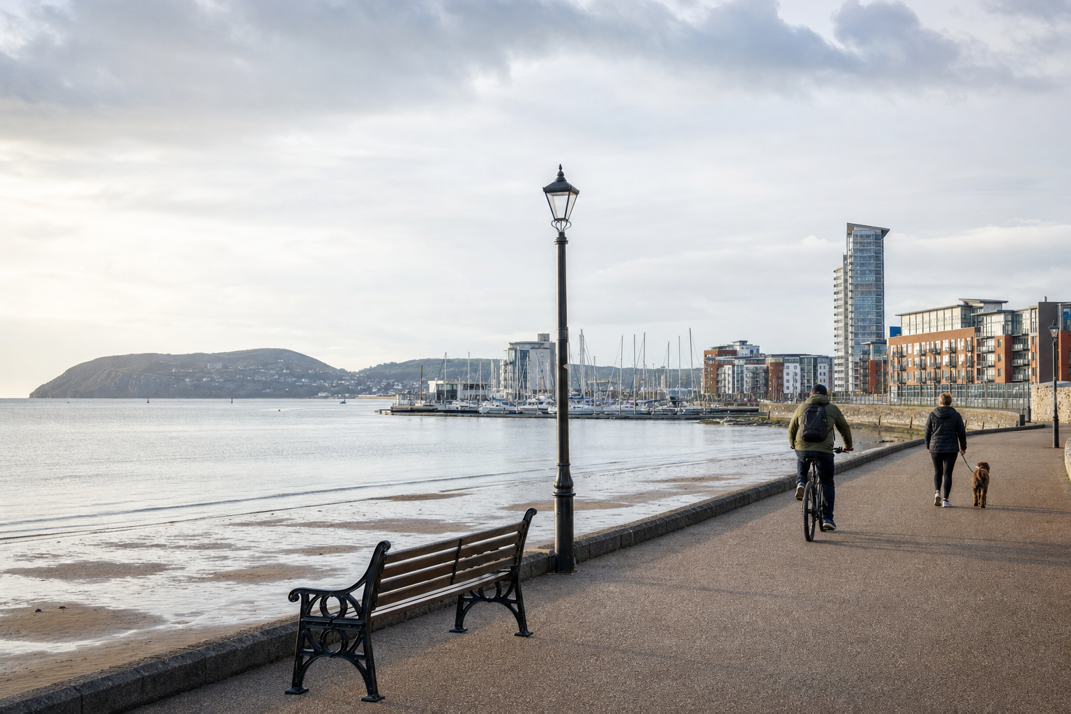A cyclist and a person walking a dog along a seaside promenade with distant buildings and a hill.