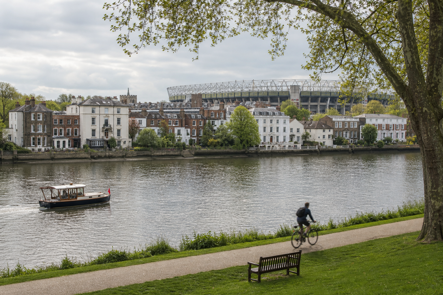 Cyclist on riverside path with a boat on the river and a stadium in the background.