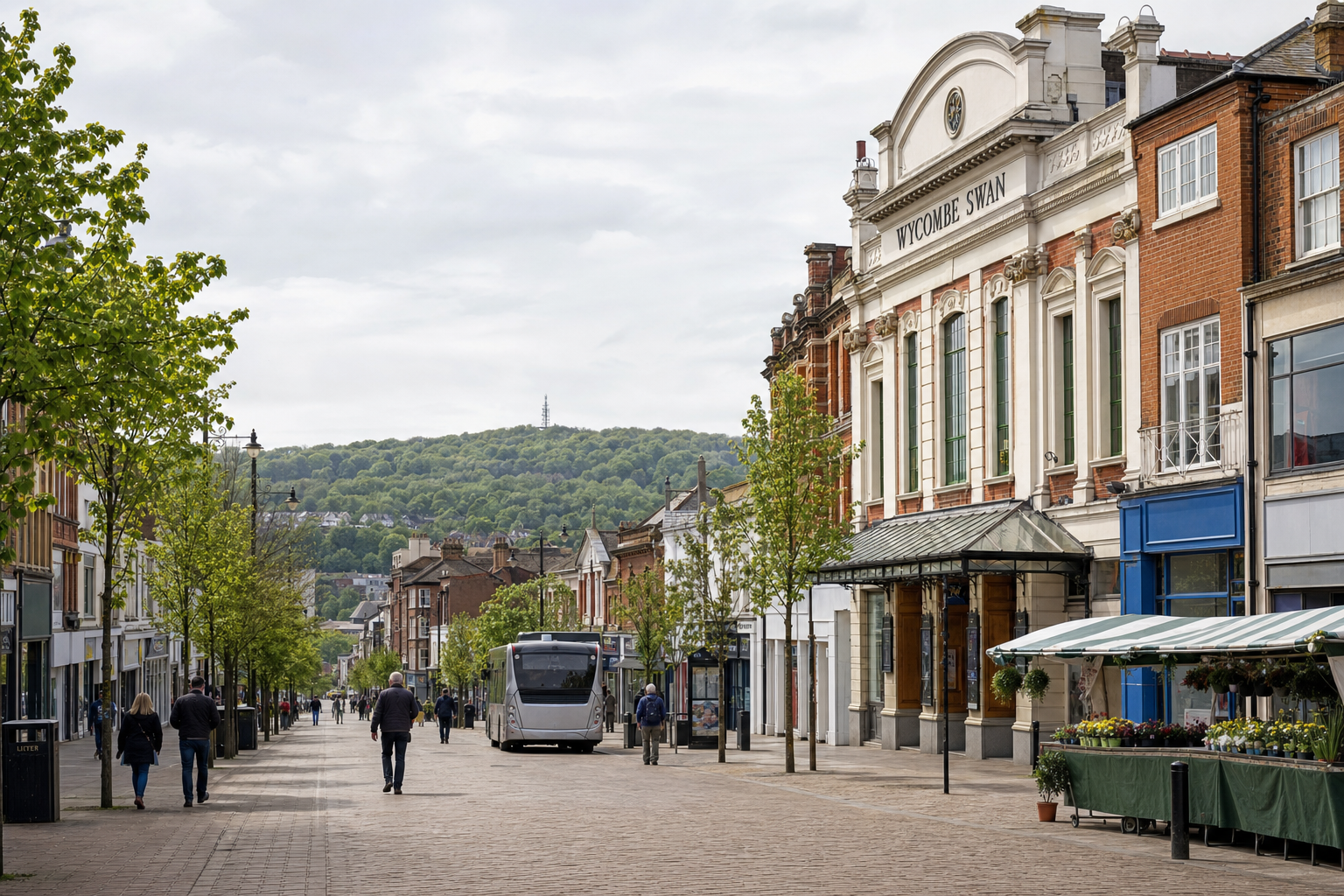 Pedestrian street in Wycombe with shops, trees, and a bus, set against a backdrop of green hills.