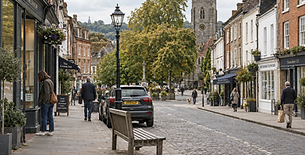A quaint cobblestone street in a British village with shops and a church in the background, under a cloudy sky.