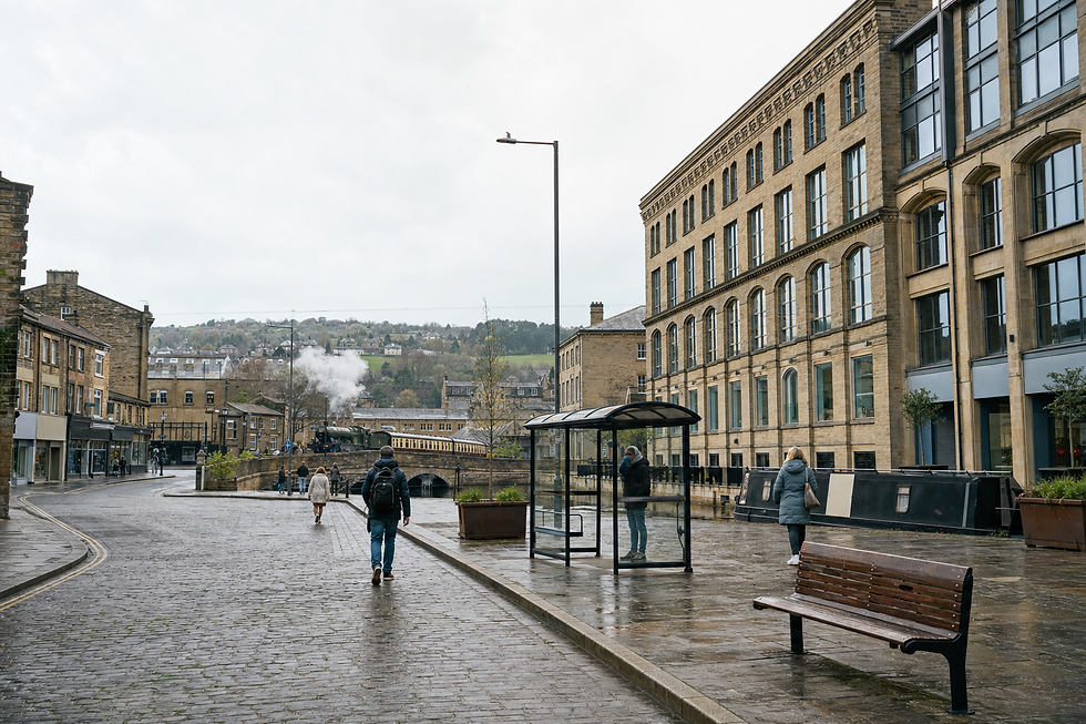 Pedestrians walk along a wet cobblestone street near historic buildings and a steam train in a quaint town setting.