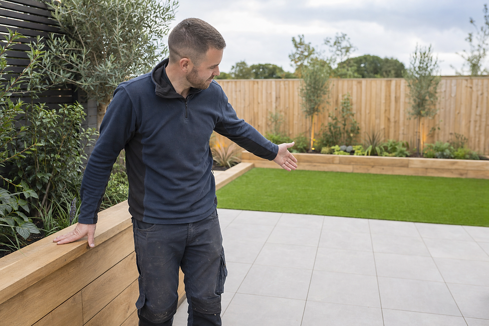 A man gestures towards a modern garden with grass, plants, and wooden fencing.