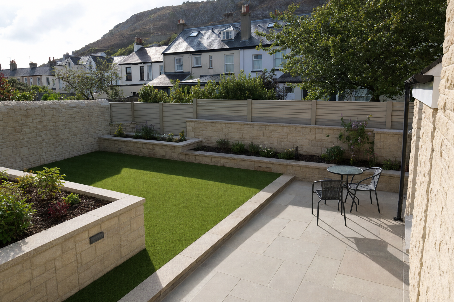 A well-maintained courtyard garden with artificial grass, stone walls, and a small patio set, surrounded by houses and a hill.