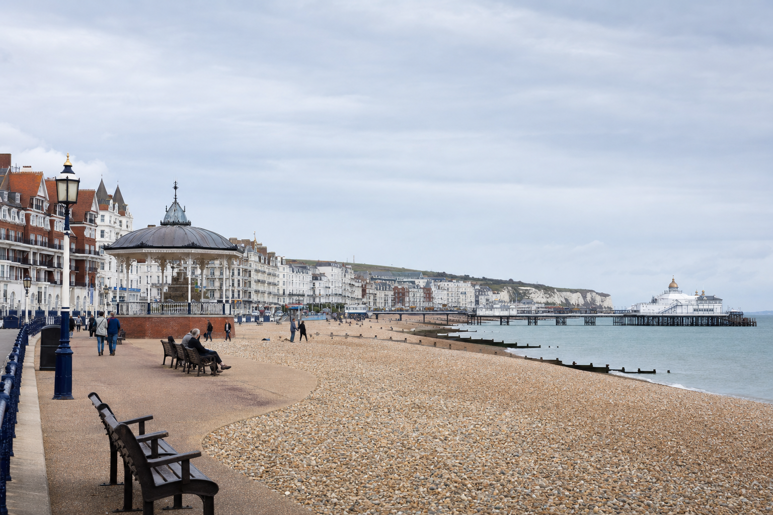 A pebble beach with a Victorian bandstand, promenade benches, and a view of a pier and coastal buildings under a cloudy sky.