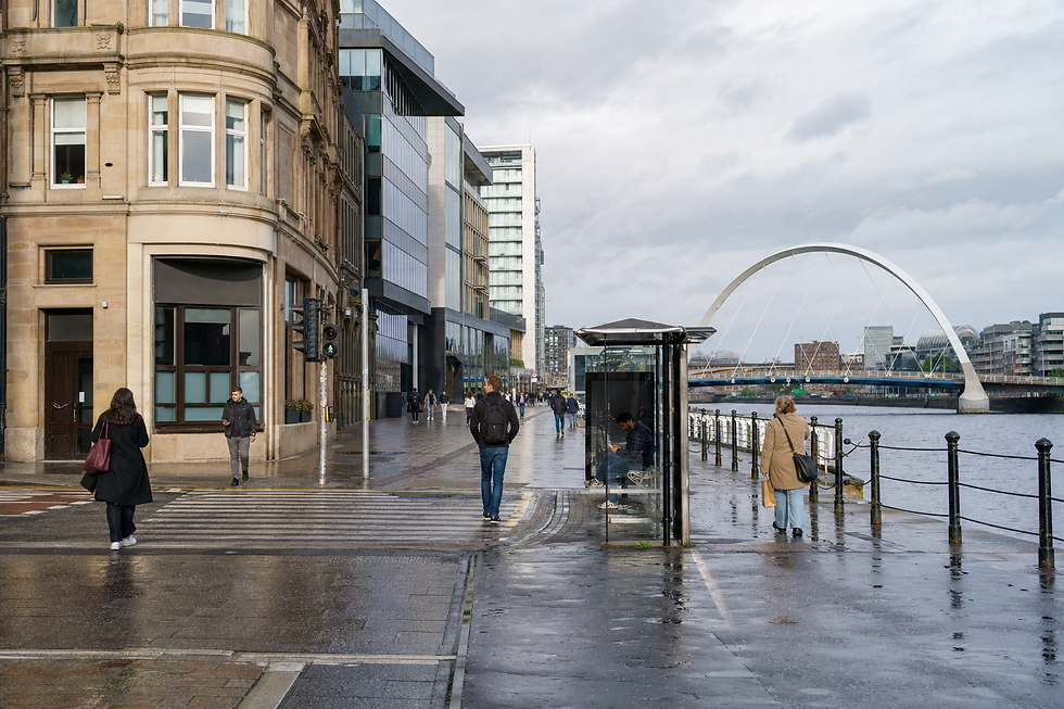 People walking along a wet riverside street lined with modern and historical buildings and a bridge in the background.