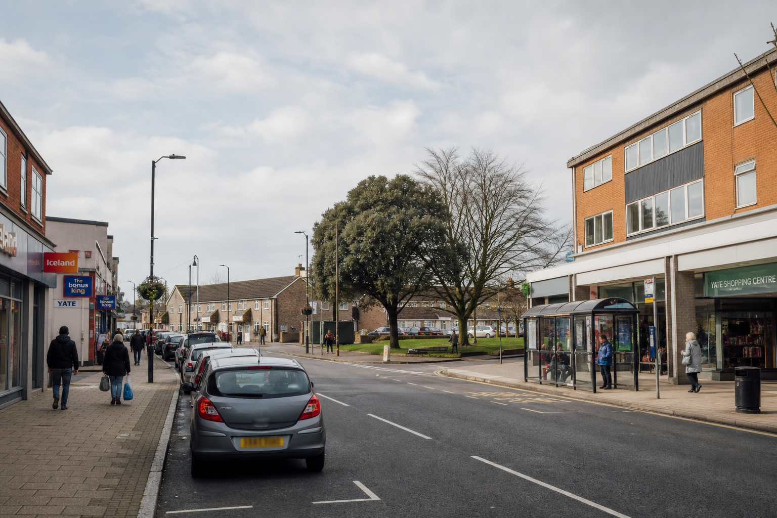 High street in Yate featuring retail shops, parked cars, pedestrians, and a bus shelter under a partly cloudy sky.